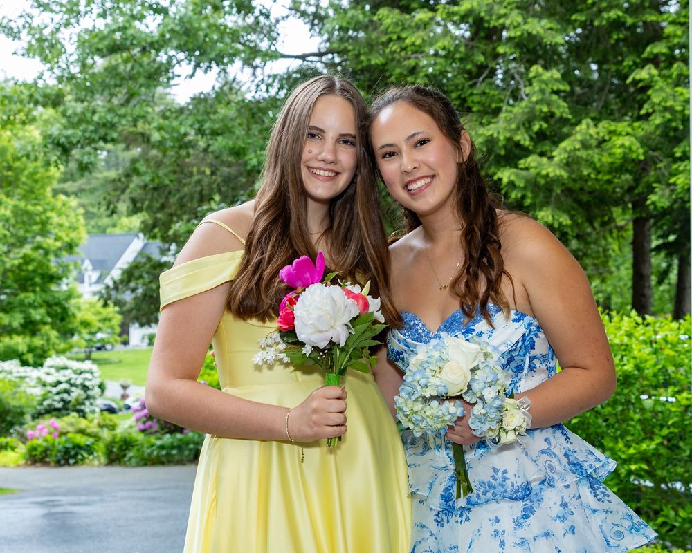 Two young women in formal dresses hold bouquets while posing together in a garden setting.