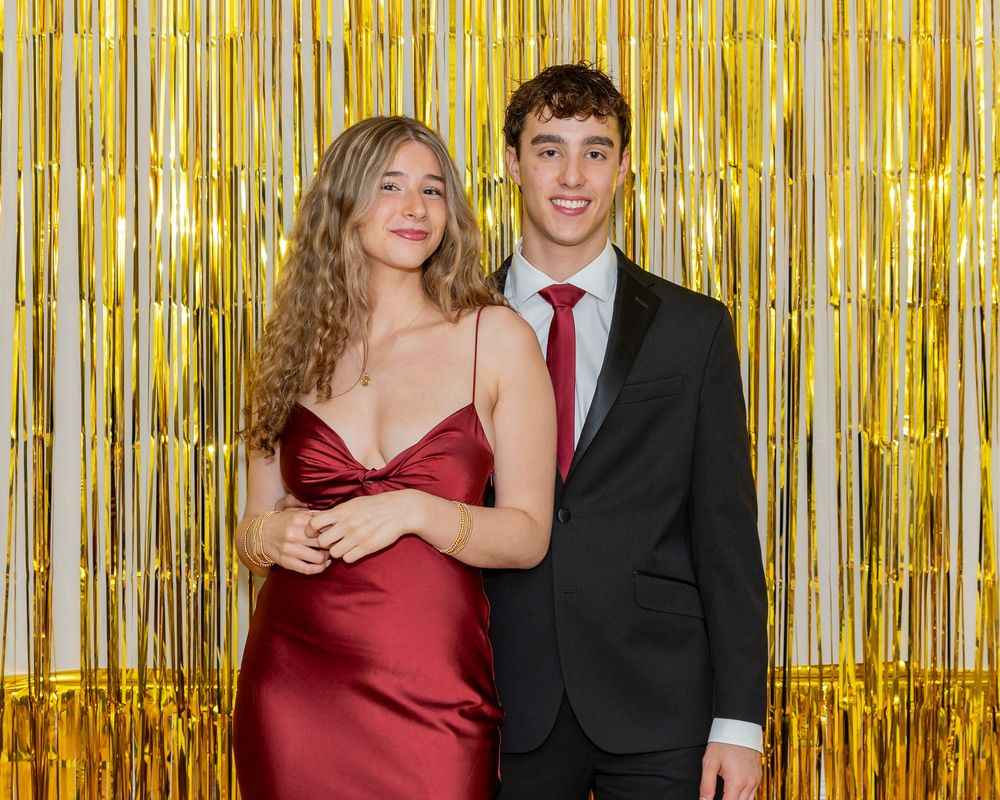 A couple poses together at a formal event against a gold metallic streamer backdrop.