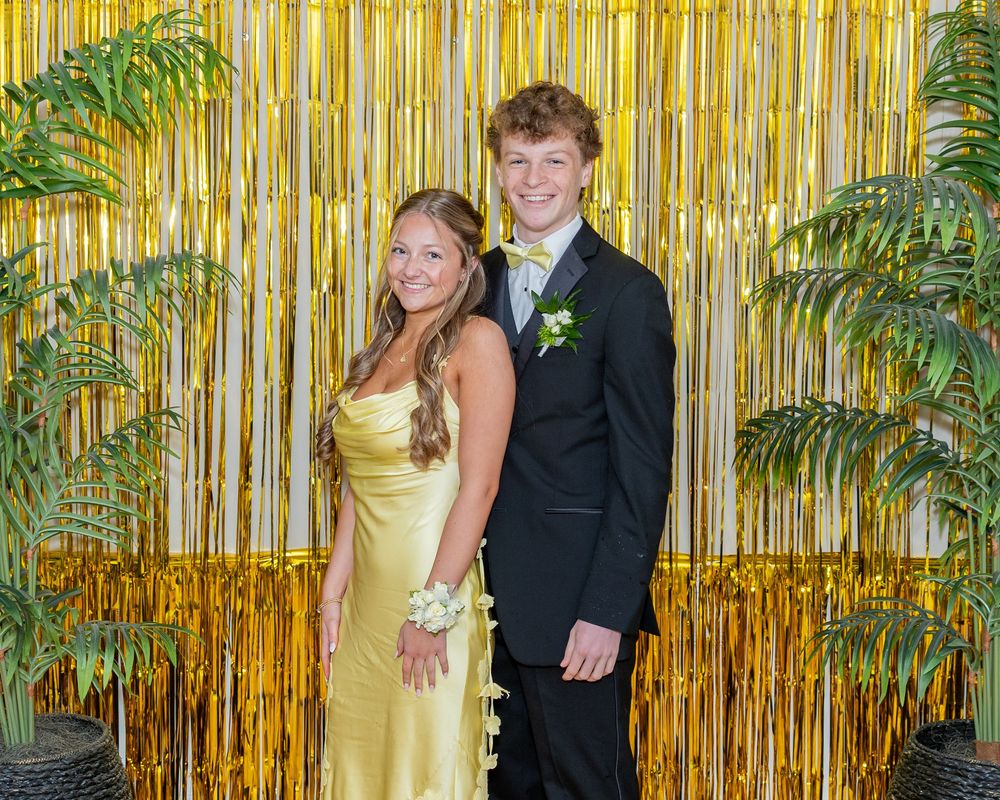 A couple poses in formal attire at a dance in front of a gold metallic backdrop with tropical plants.