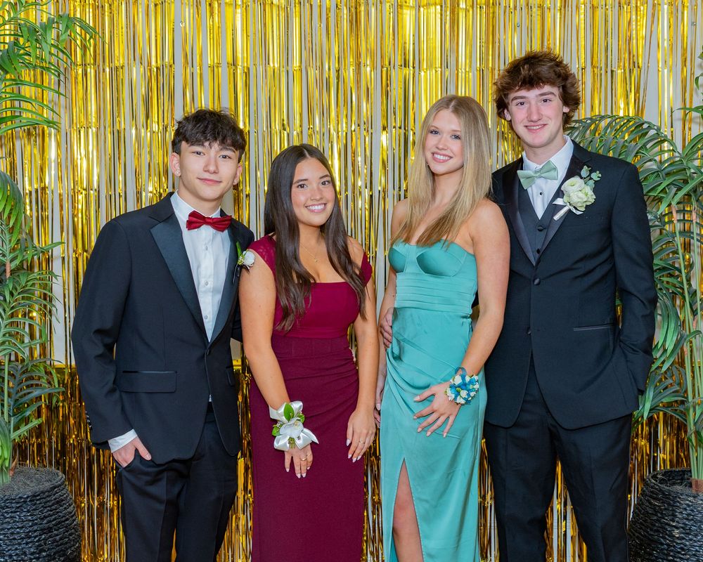 Group poses at formal dance event in front of shimmering gold backdrop wearing elegant dresses and suits.