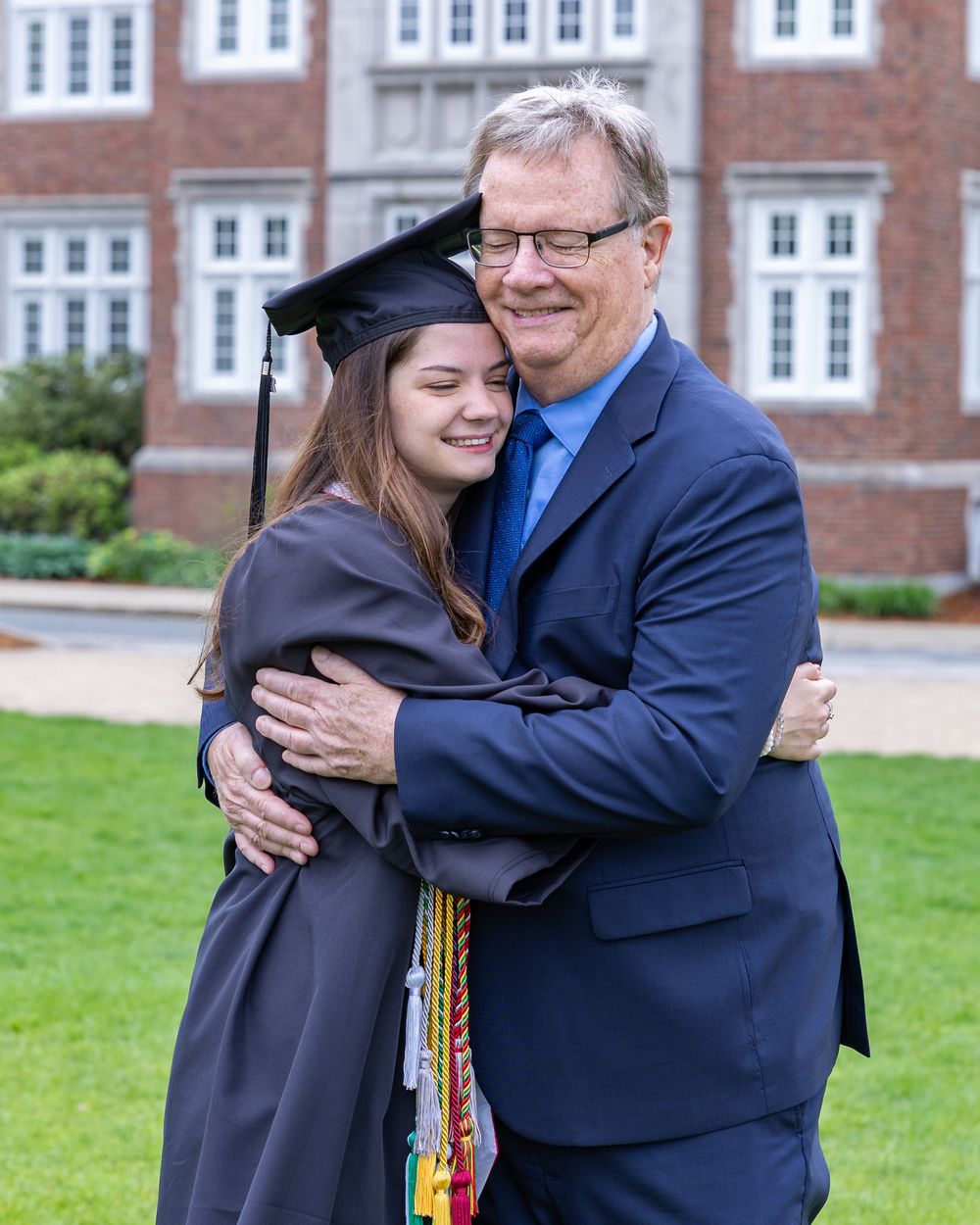 A graduate in cap and gown embraces a companion in a blue suit standing on a lawn in front of a brick building.