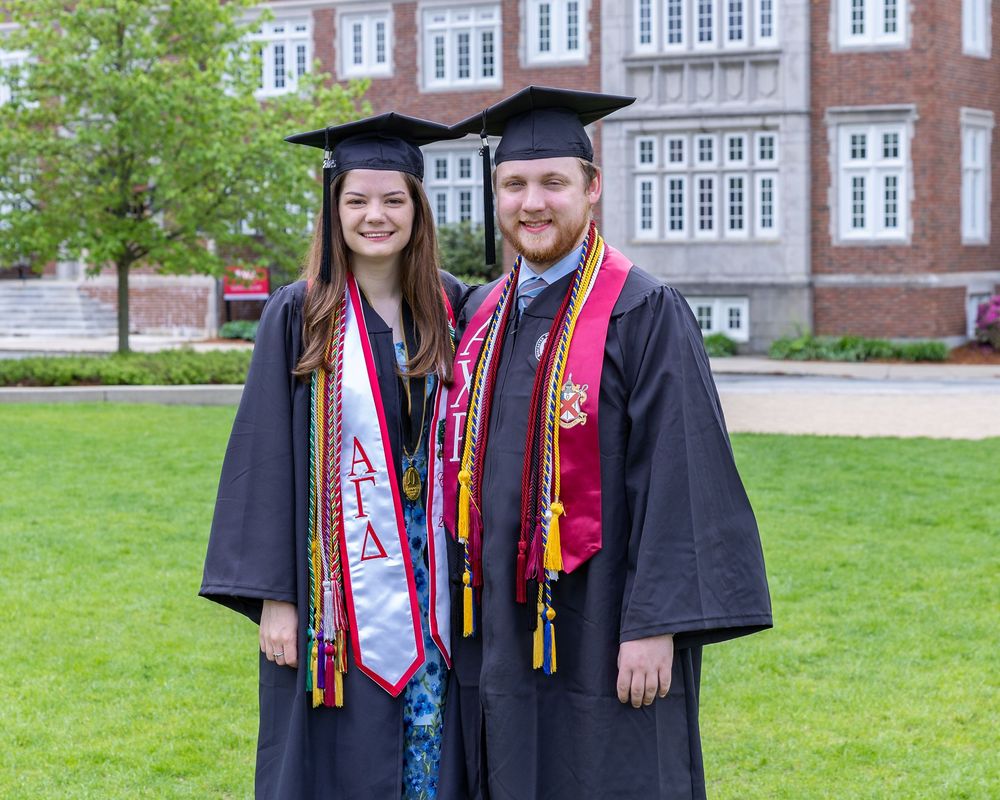 Two graduates in black caps and gowns with honor cords and sashes pose on a grassy lawn in front of a brick building.