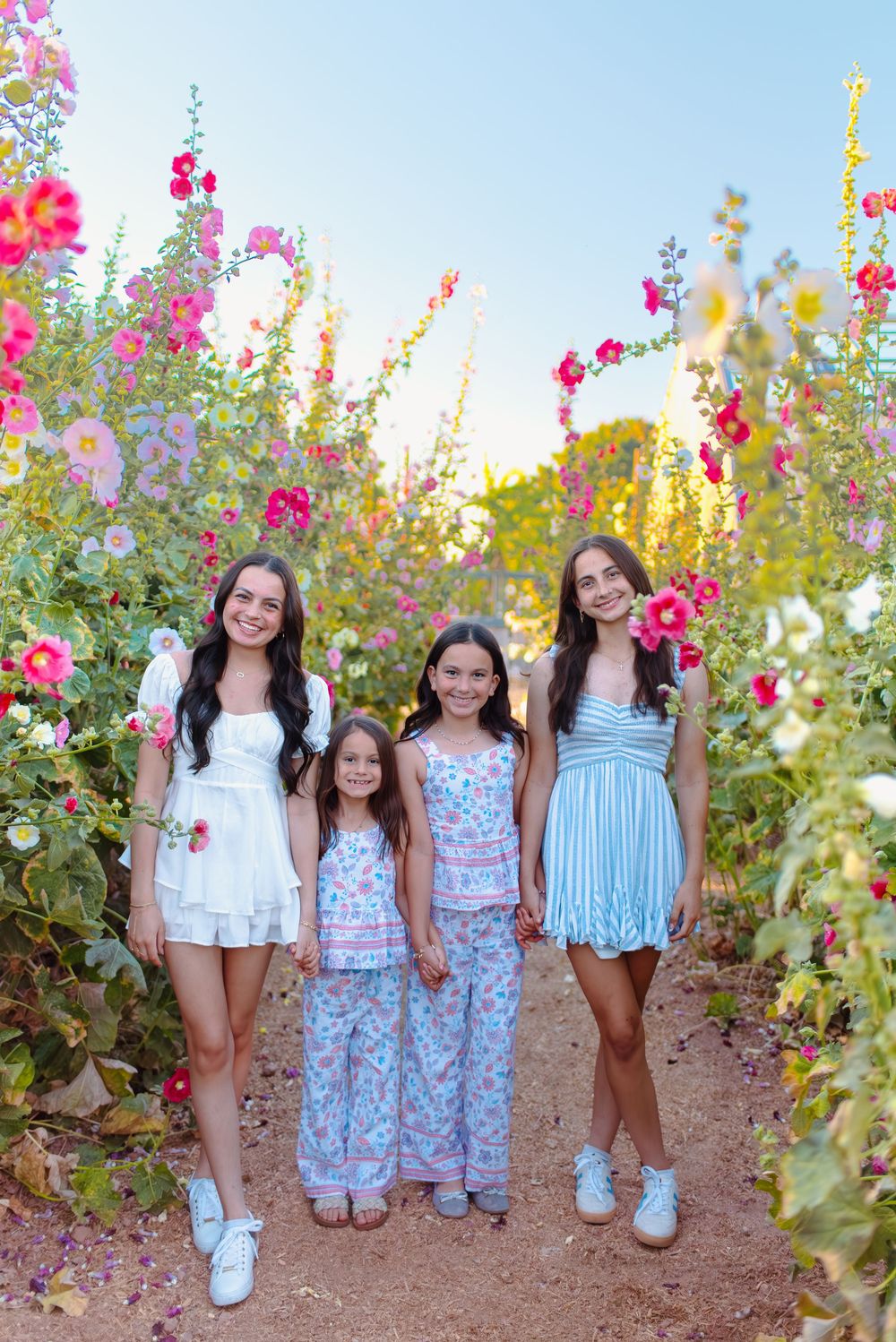 Four people in spring dresses pose together by bright pink hollyhock flowers in a garden path during golden hour.