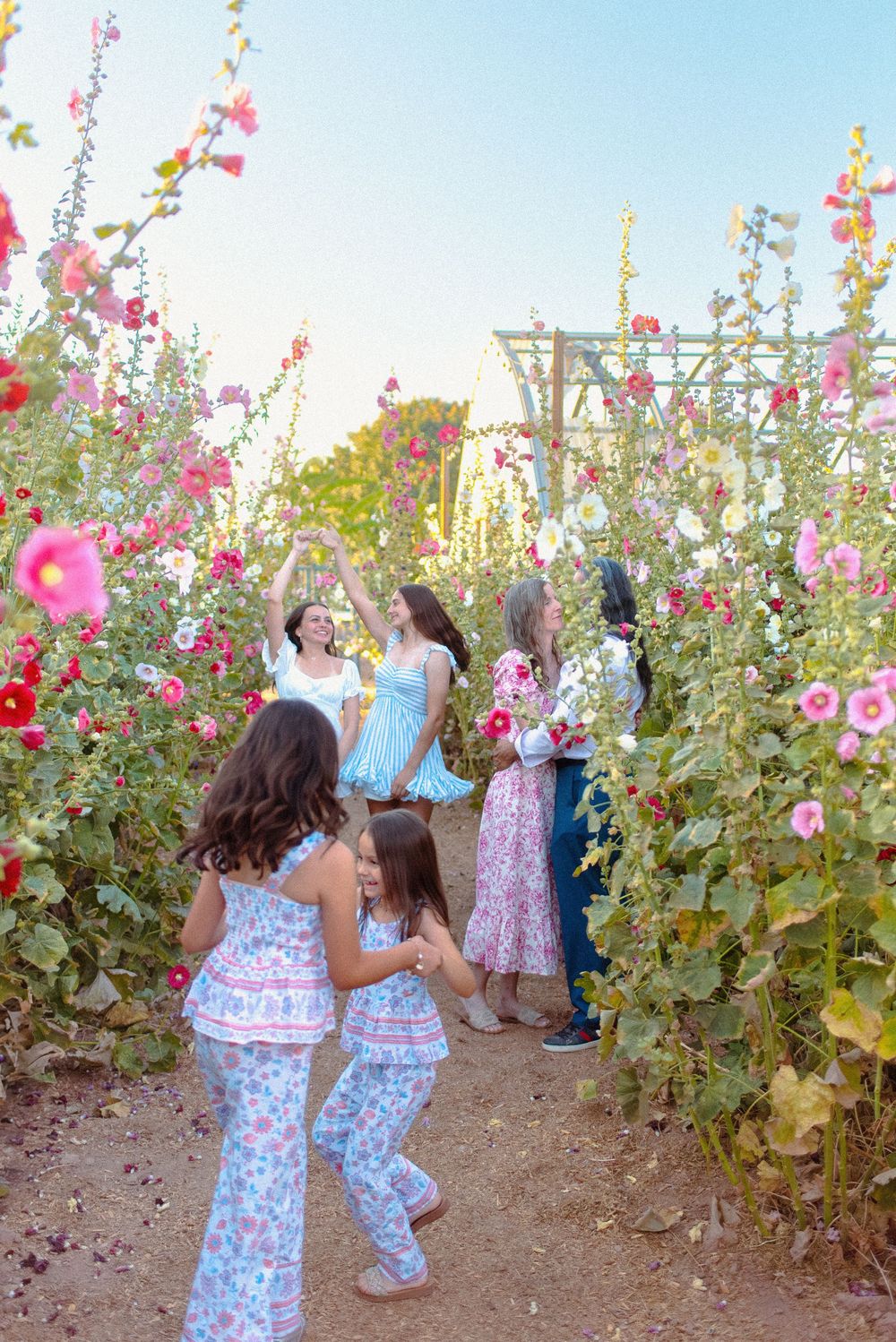 Children dance and play among towering pink hollyhock flowers in a spring garden at sunset.