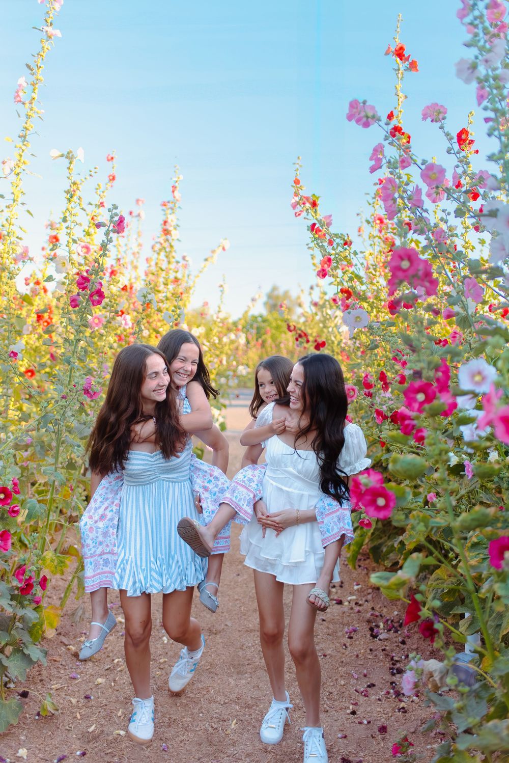 Four people in white outfits laugh and dance in a Arizona flower garden with vibrant pink hollyhock flowers blooming around them.