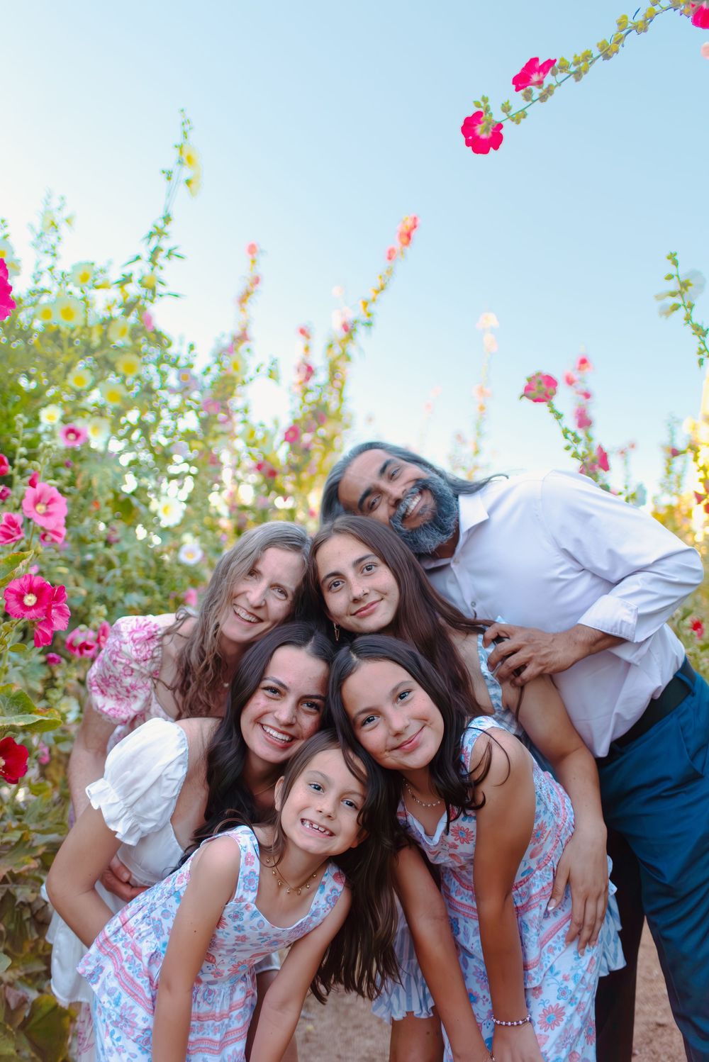 A happy family poses together outdoors among blooming pink hollyhock flowers on a sunny day.
