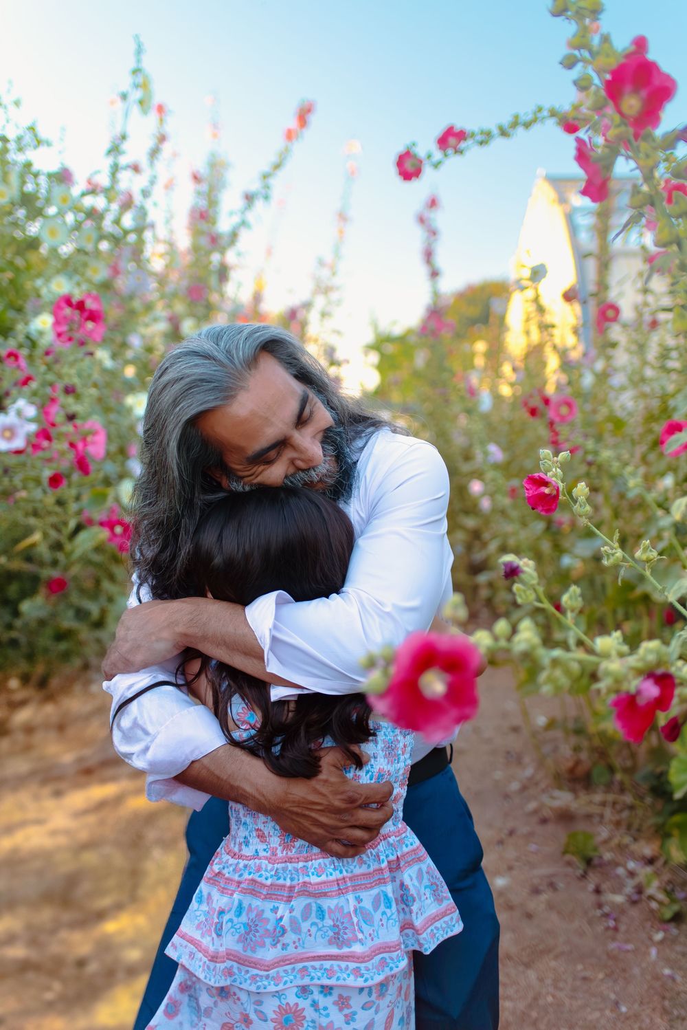 A tender embrace between two people among blooming pink hollyhock flowers in a garden at sunset.