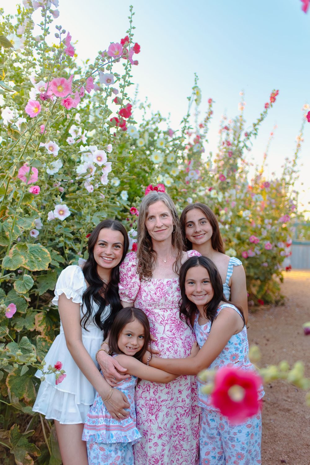 A family poses together among pink hollyhock flowers in an outdoor garden setting.