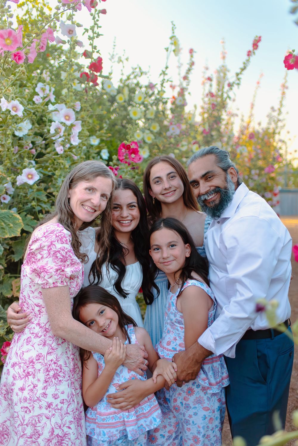 A family poses together in front of blooming pink and white hollyhock flowers in a garden during sunset.