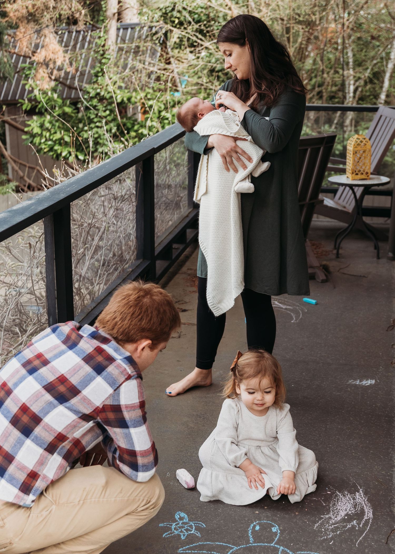A family spends time together on a porch as a child draws with chalk on the ground while others look on.