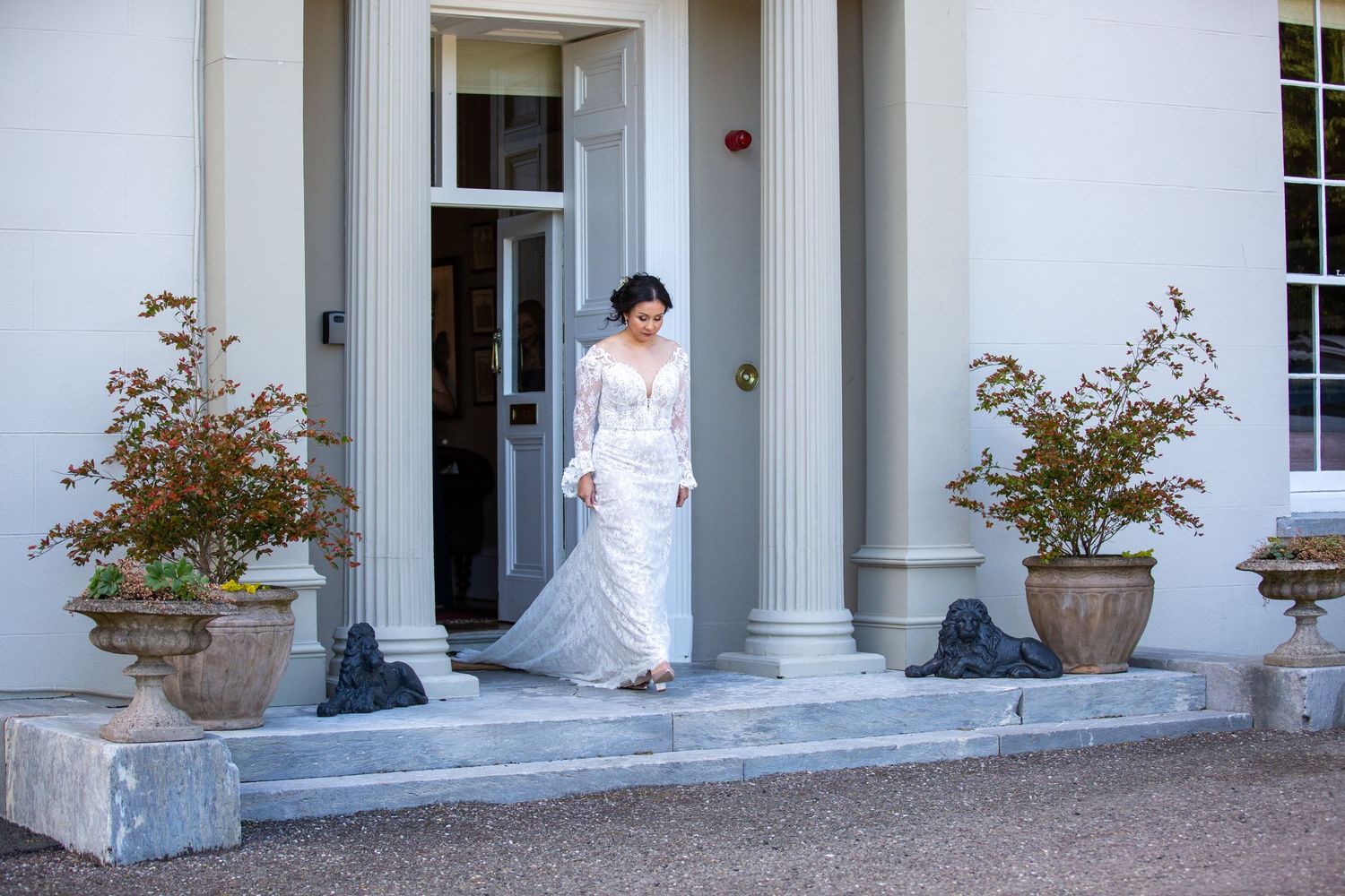 A bride in a white lace gown stands elegantly on the steps of a classic columned entrance with potted plants.