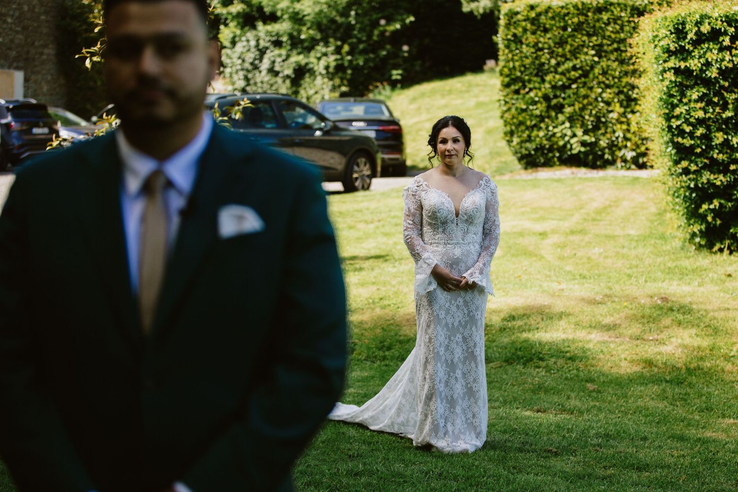 A bride in a lace gown waits on a grassy lawn while someone stands in a suit in the foreground.