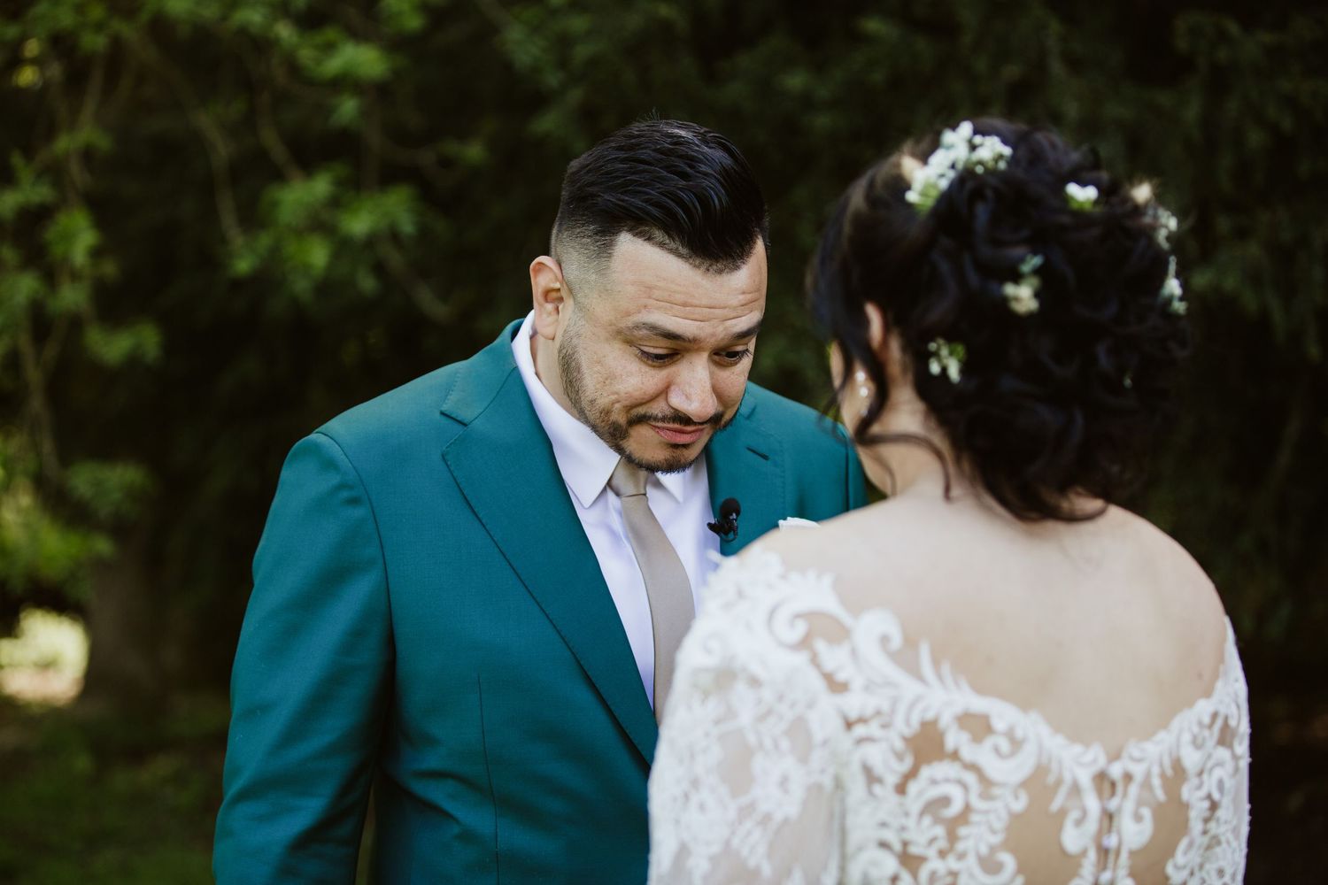 A couple in wedding attire share an intimate moment outdoors, with the groom wearing a teal suit jacket.