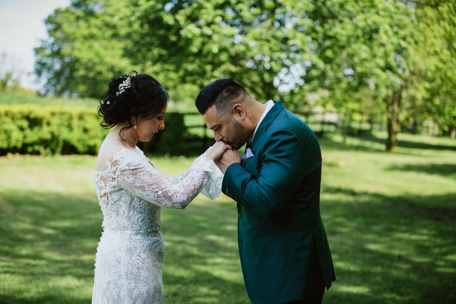 A couple shares an intimate moment in a garden setting with the person in teal suit kissing their partner's hand.