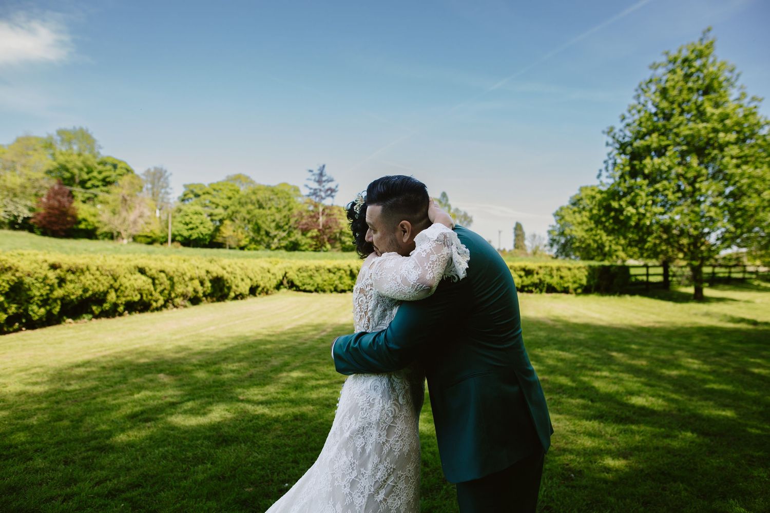 A couple embraces on their wedding day in a lush garden with green trees and a bright blue sky.