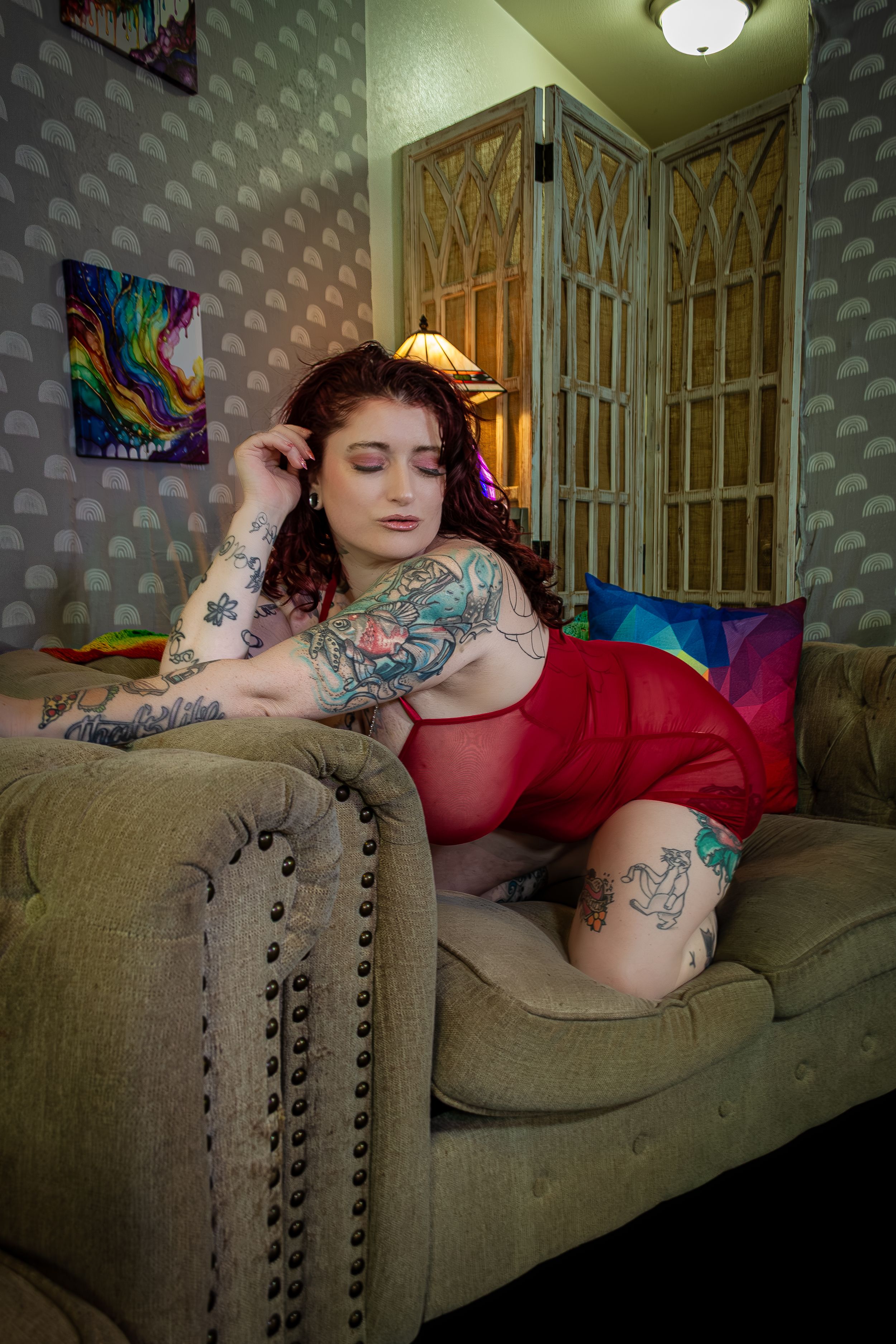 Woman kneeling on on a tan couch wearing red lingerie, with visible tattoos and colorful art on the wall.