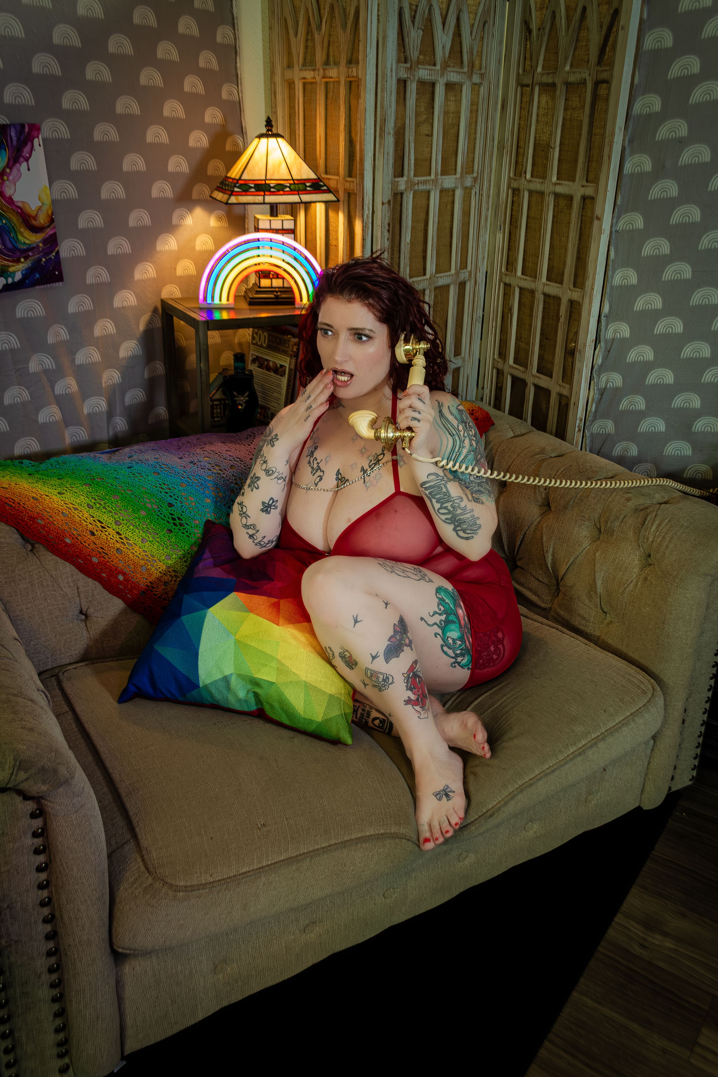 A retro-styled boudoir photo with rainbow pillows, vintage phone, and warm lamp lighting on a beige couch.