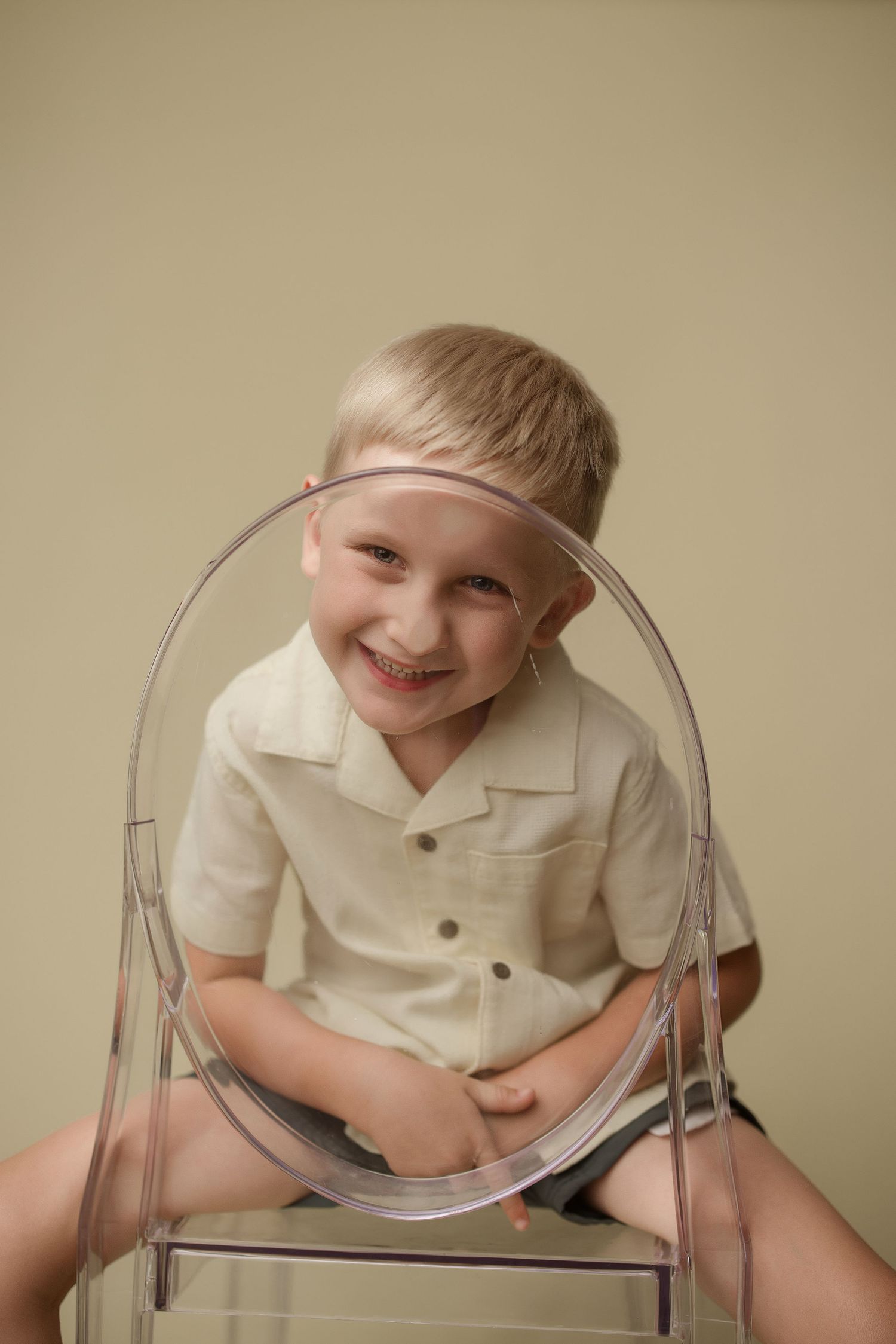 Child wearing a white button-up shirt sits on a transparent acrylic chair with a joyful expression against beige background.