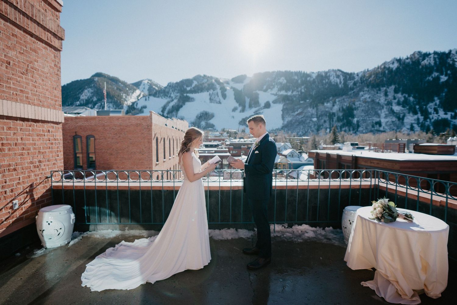 Two people in wedding attire stand together on a rooftop terrace of Hotel Jerome with snow-capped mountains in Aspen during winter.
