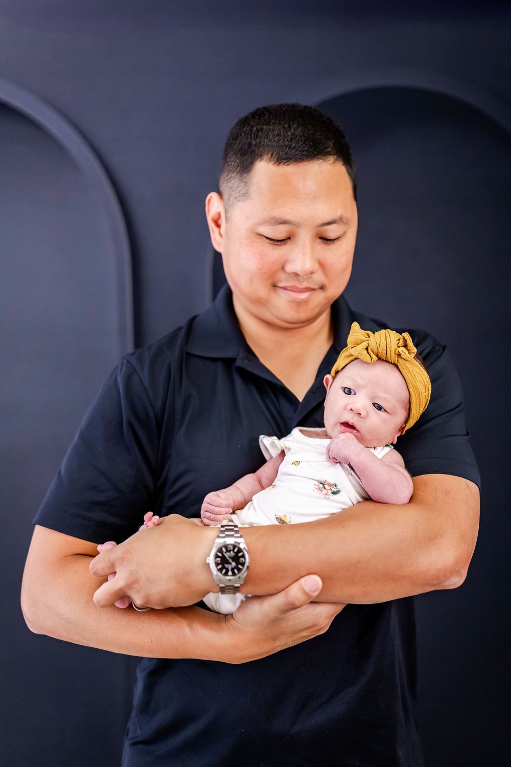 Sweet portrait series showing an infant wearing a yellow bow headband being cradled in arms against a dark backdrop.