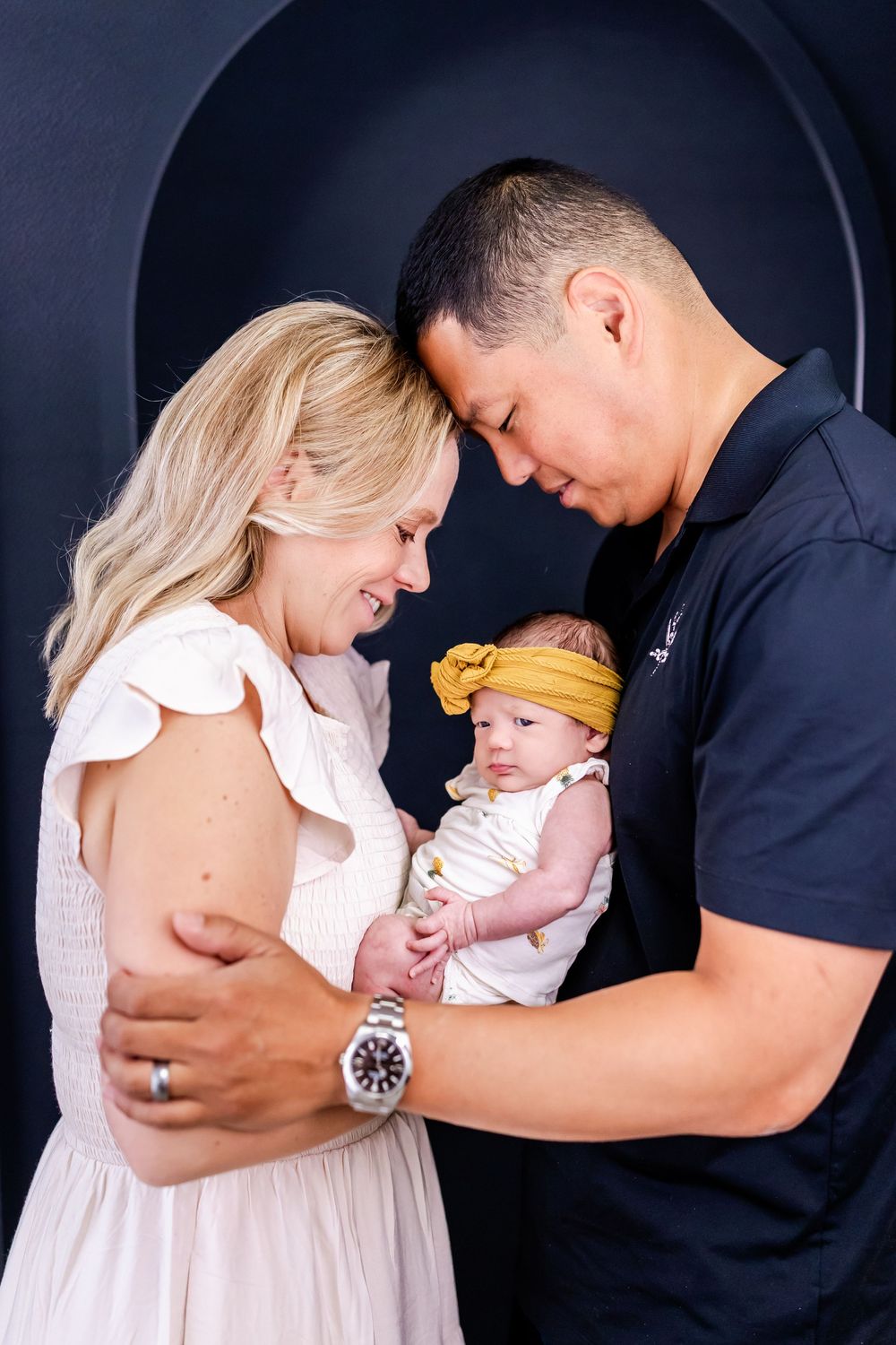 A couple shares a tender moment with their newborn baby against a dark background.