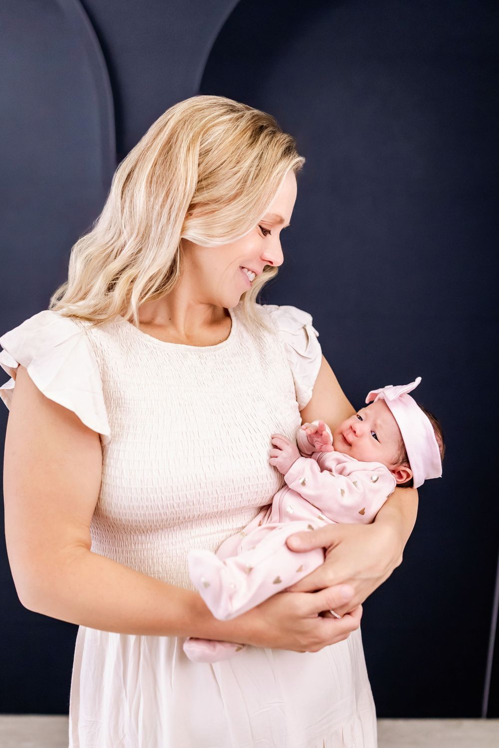 A woman in a white dress holds a newborn baby in her arms against a dark backdrop in a studio portrait session.