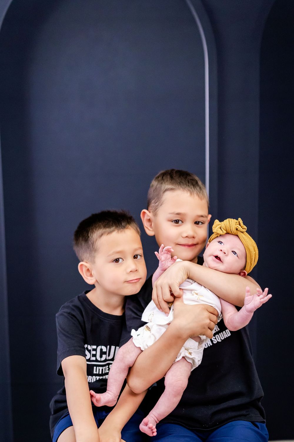 Two young children in dark shirts hold and interact with a baby wearing yellow on a dark background.