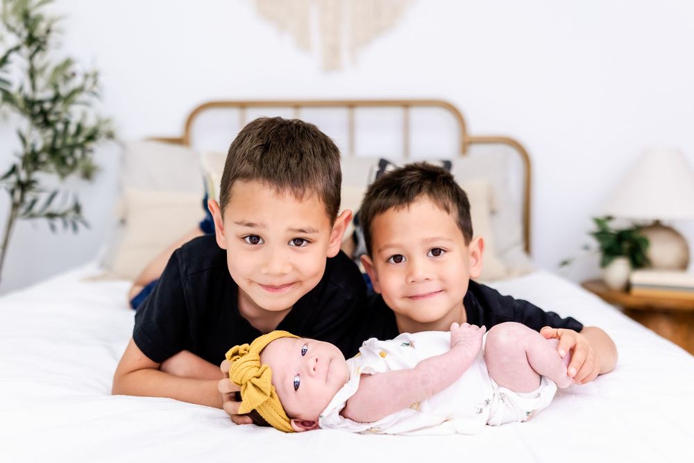 Two children laying on bed with baby dolls at home with white bedding and plants in background.
