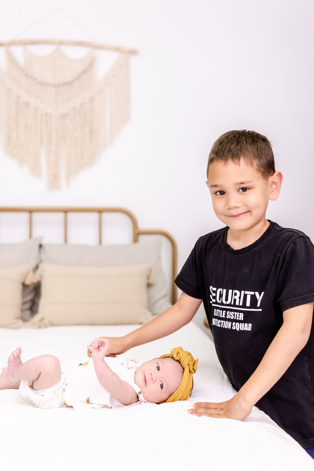 Two children on a bed with one wearing a black security shirt and decorative wall art in the background.