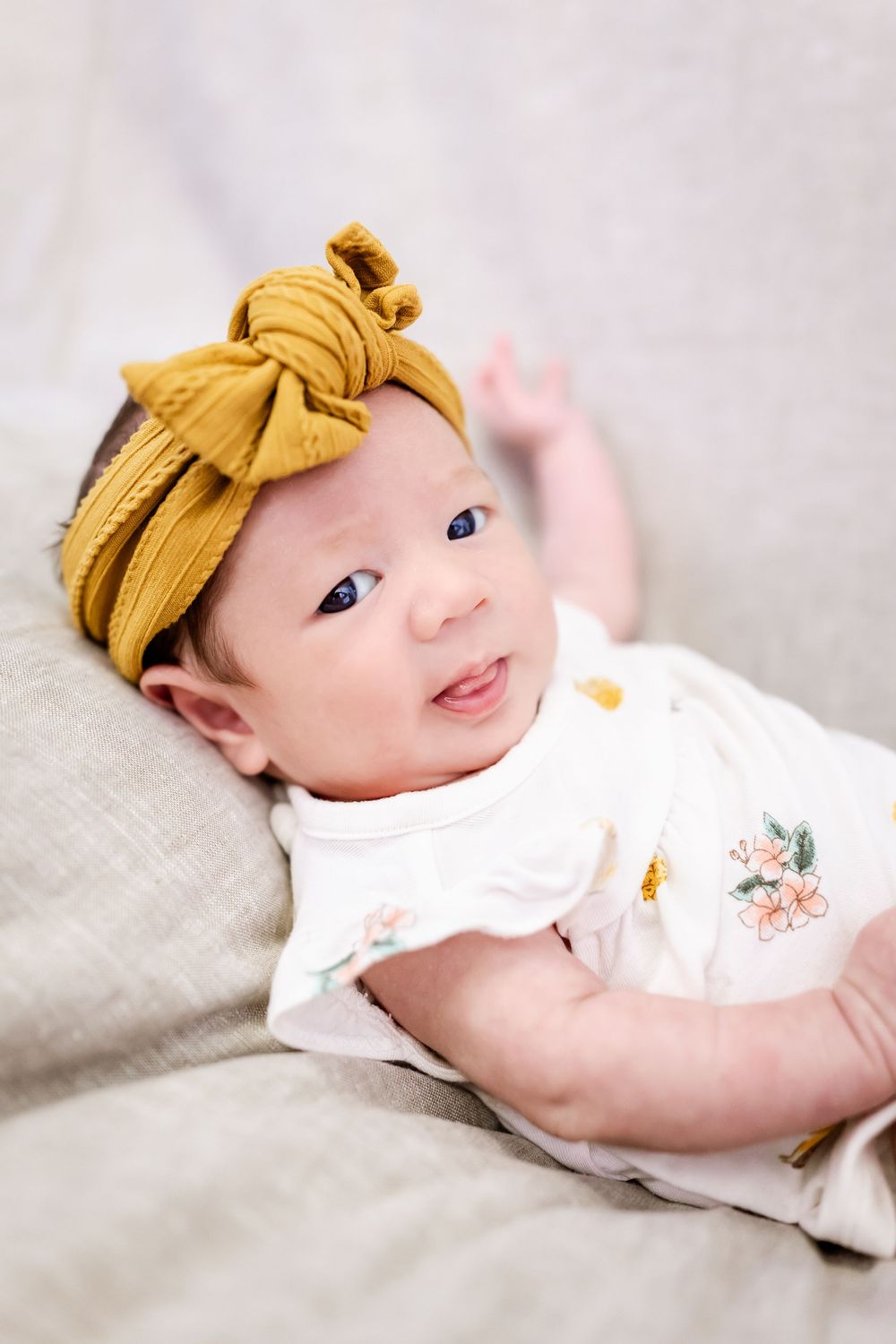 Adorable baby wearing white outfit and mustard yellow bow relaxing on soft gray bedding.