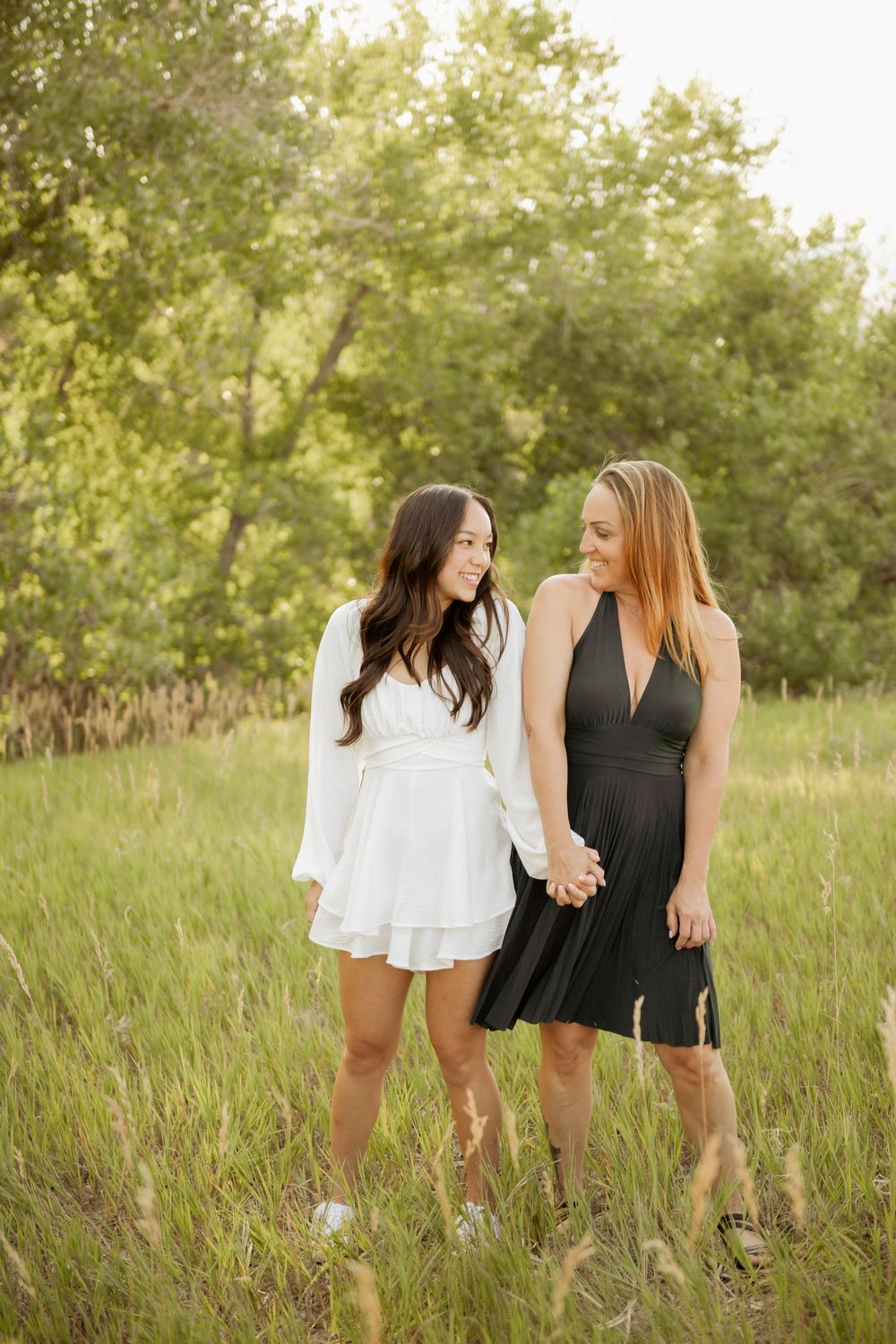 Two figures in contrasting white and black dresses walk together through a sunlit meadow on a summer afternoon.