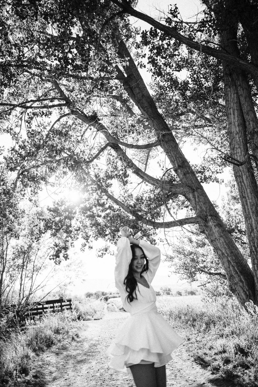 A woman in a white dress poses under a canopy of trees along a sunlit rural path in black and white photography.
