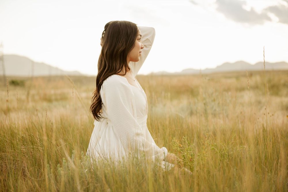 A figure in a flowing white dress stands gracefully in a golden field of tall grass at sunset.