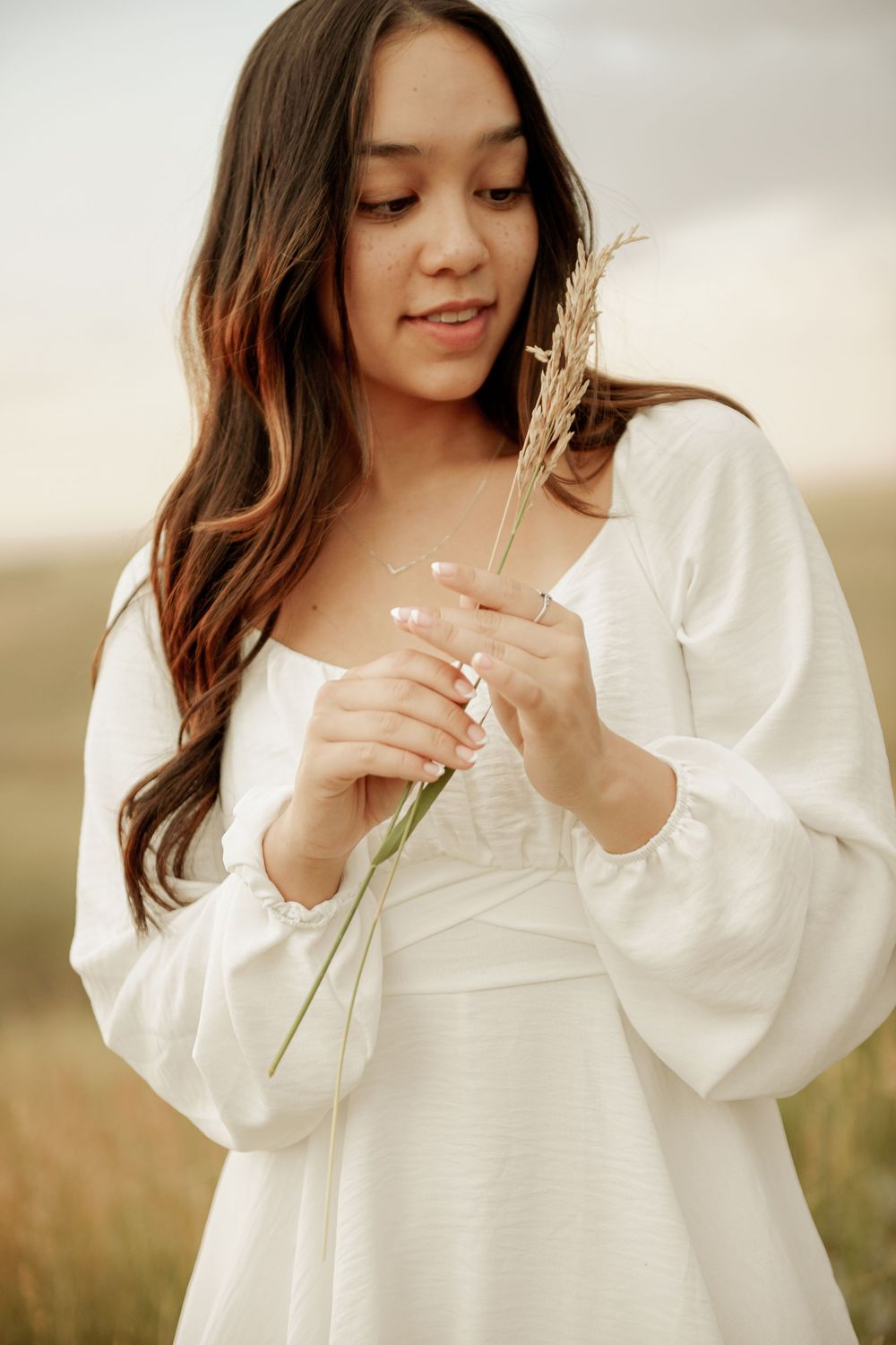 Person in flowy white dress holds dried wheat stalk while standing in golden field during sunset.