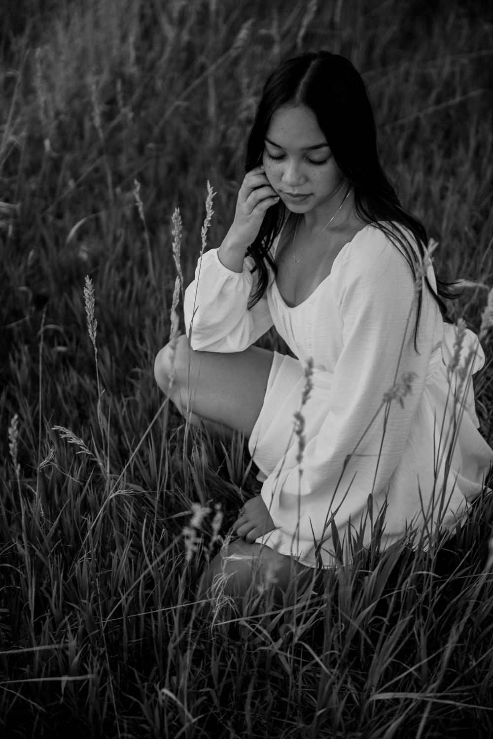Black and white artistic series of a person in a white dress sitting contemplatively among tall grass in a field.