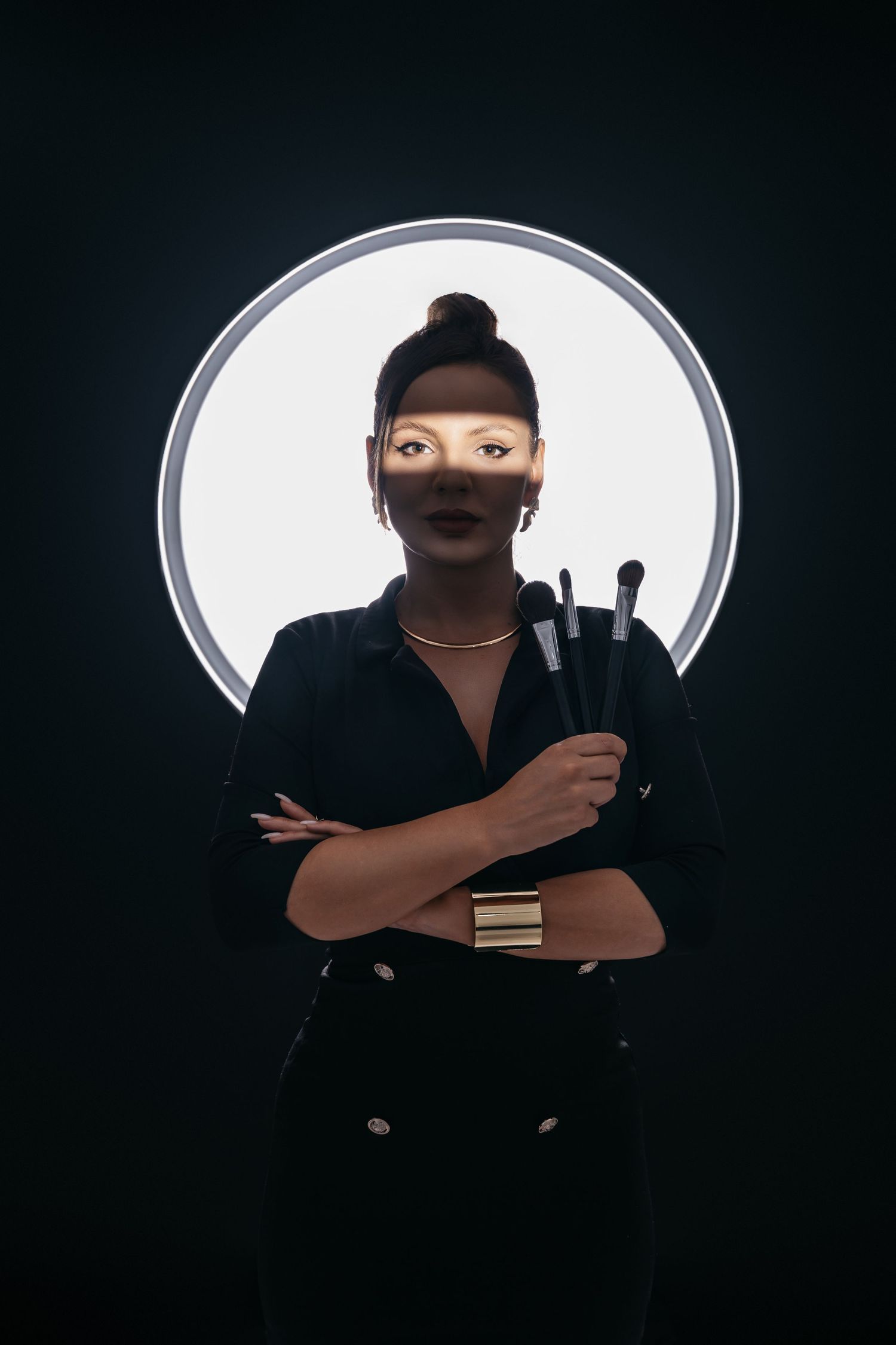 Artistic beauty portrait with circular white background featuring figure in black holding makeup brushes against dark surround.
