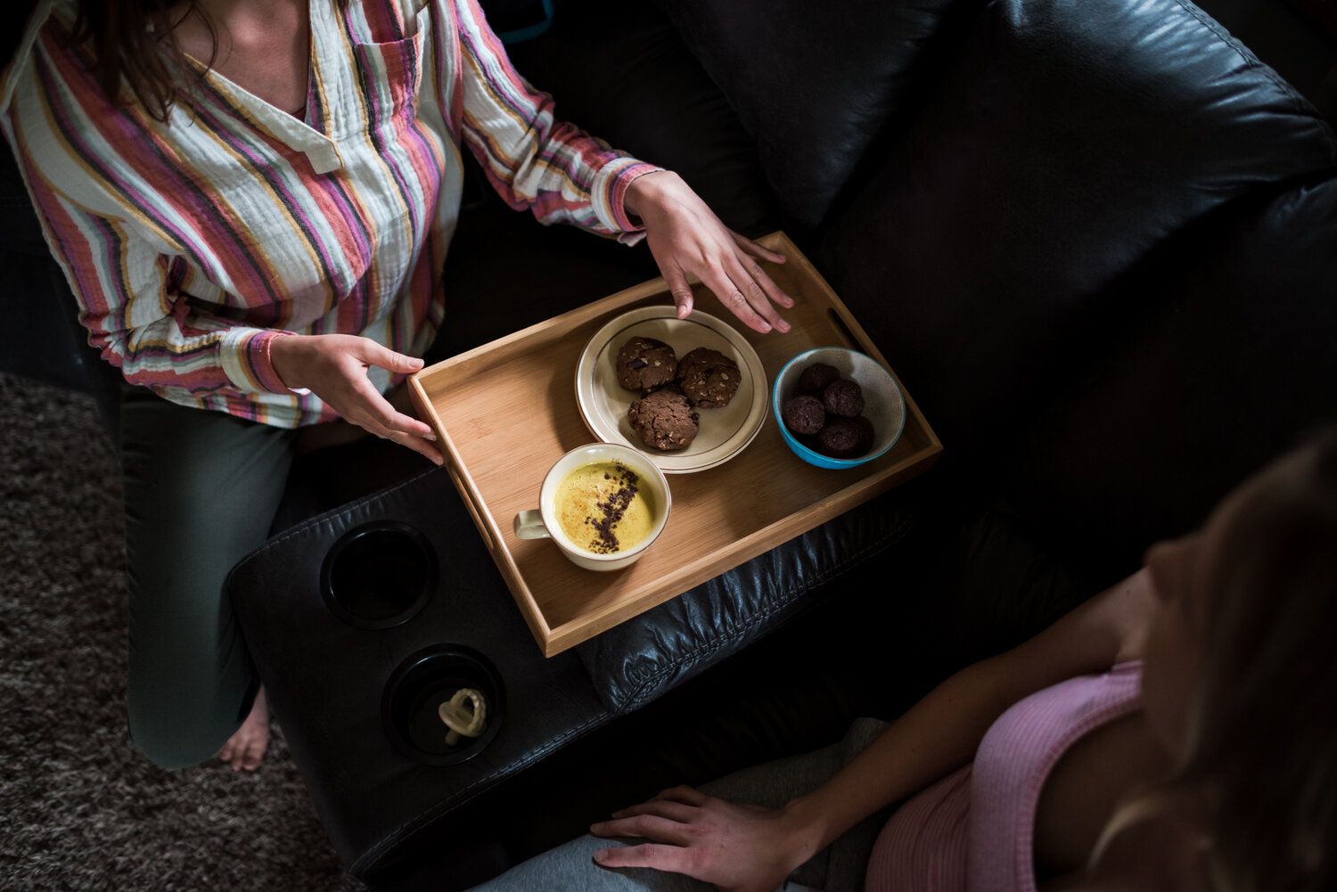 Overhead view of wooden serving tray with food and drinks on dark couch during cozy indoor meal.
