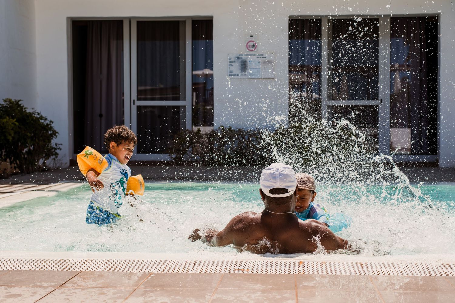 a father and his children splash and play in a swimming pool on a sunny day, creating dramatic water sprays and ripples.