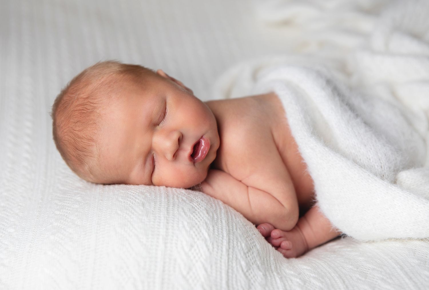 A sleeping newborn baby wrapped in soft white blankets rests peacefully on textured white bedding. During a London baby photoshoot