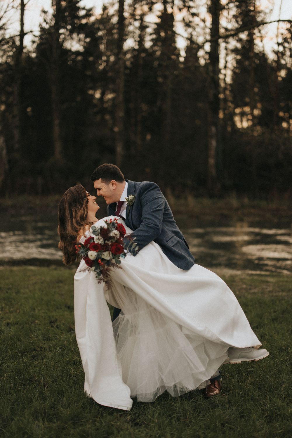 A romantic wedding portrait of a couple sharing a dip kiss in a forest setting at sunset with the bride holding a red bouquet.
