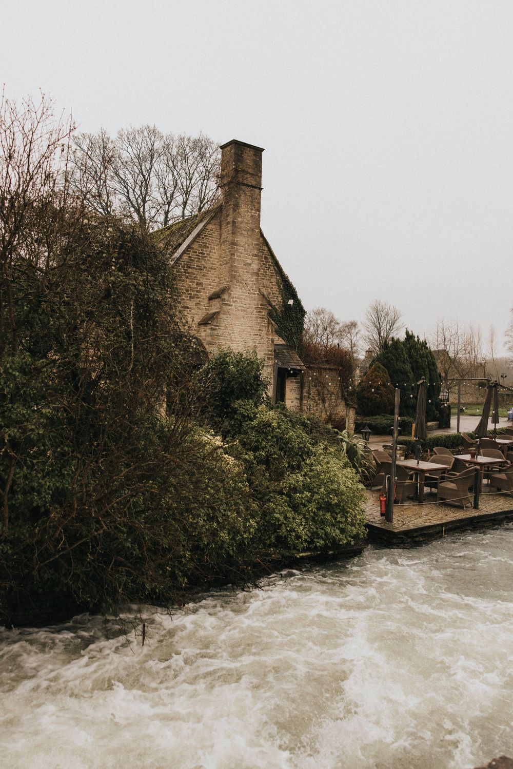 Historic stone building stands beside turbulent rushing waters during flood conditions along a riverbank.