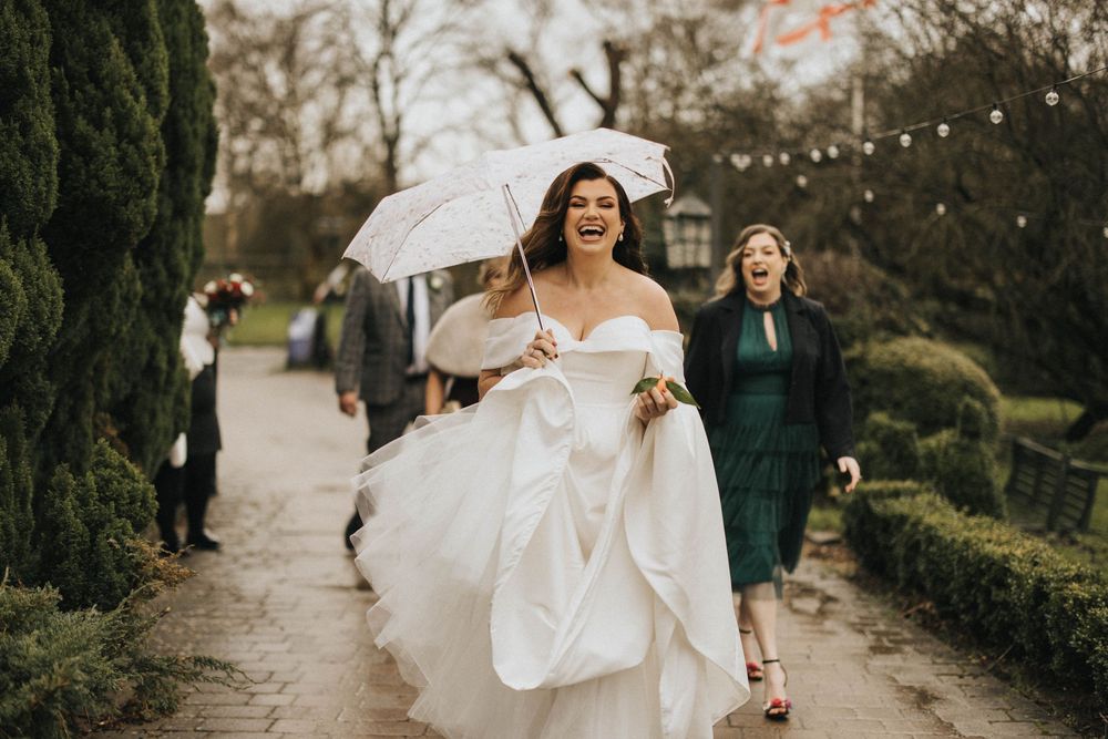 A joyful bride in a flowing white gown walks along a garden path while carrying her dress on a cloudy day.