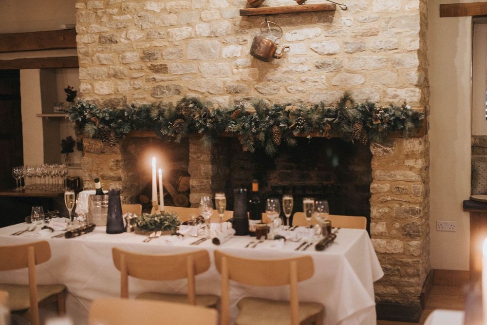 Elegant holiday dinner table setting with white tablecloth, candles and evergreen garland decorating stone wall.