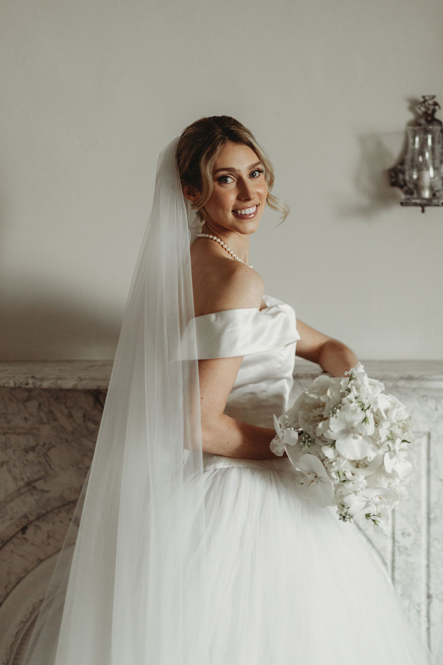 A bride in a flowing white wedding dress and veil holds an elegant white orchid bouquet while standing by a marble wall.