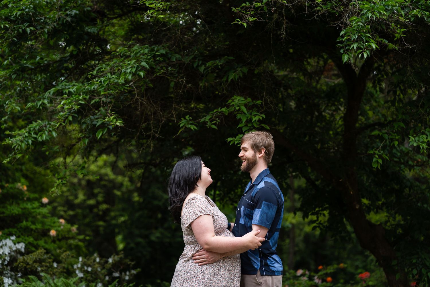 Parents smile and hug in front of tall trees at the Seattle Arboretum.