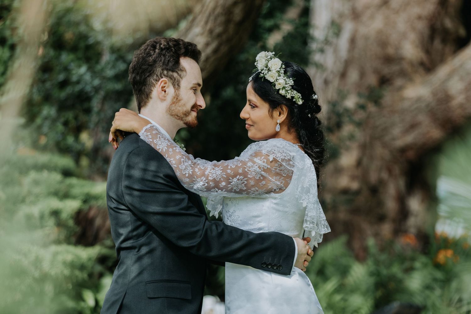 A romantic wedding couple embraces outdoors with the bride wearing a lace dress and floral crown in a garden setting.
