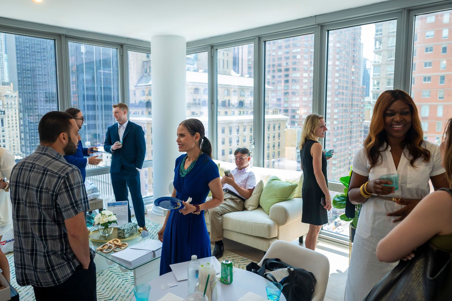 People enjoy drinks and conversation at a rooftop party with city skyline views through large windows.