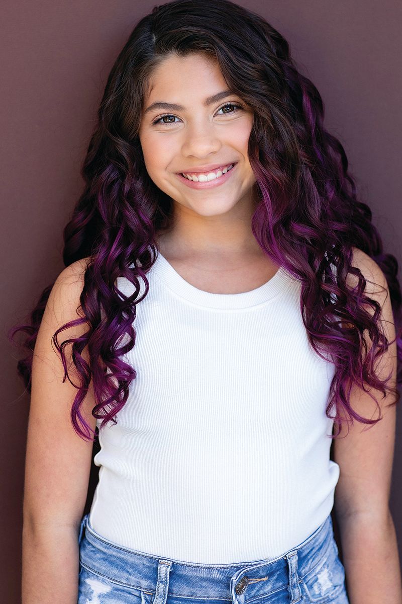 A person in a white sleeveless top showing off vibrant purple ombre curls in hair against a brown backdrop.