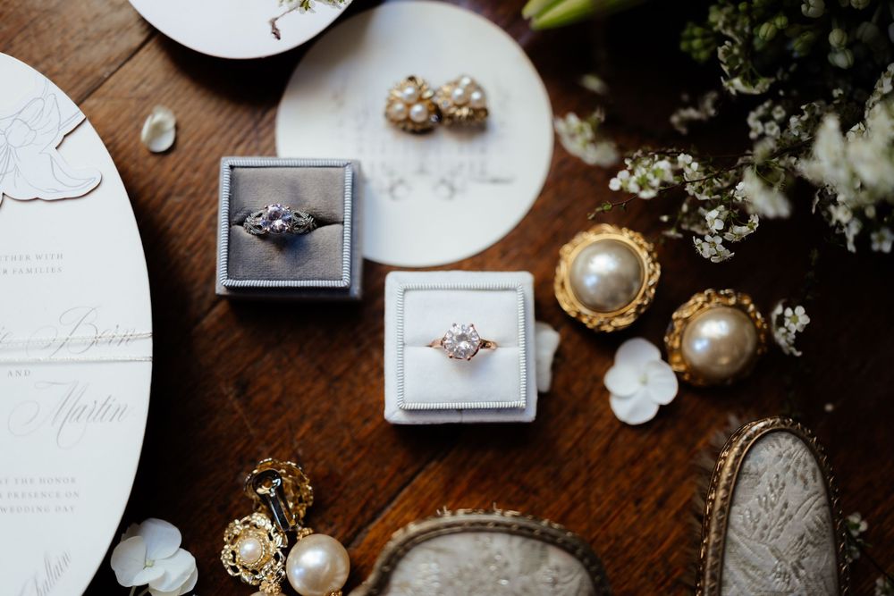 Two engagement rings in boxes sit on a wooden surface with pearl decorations and scattered white details.