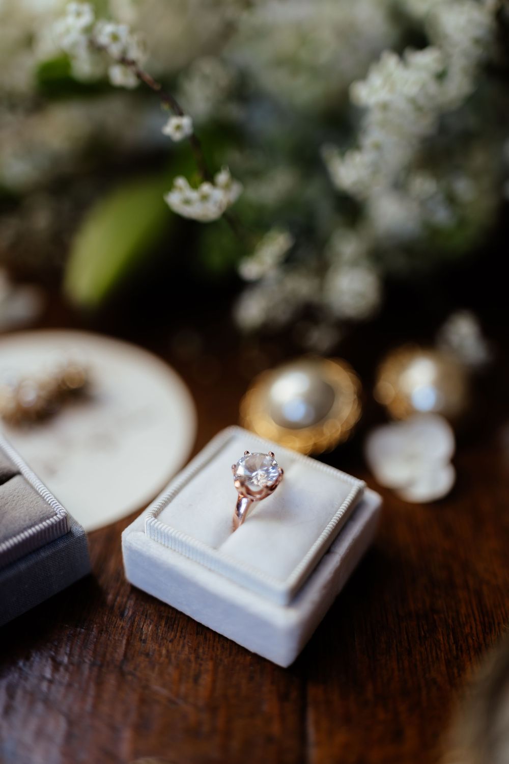 Close up of a delicate engagement ring on white fabric with soft white flowers in the background.